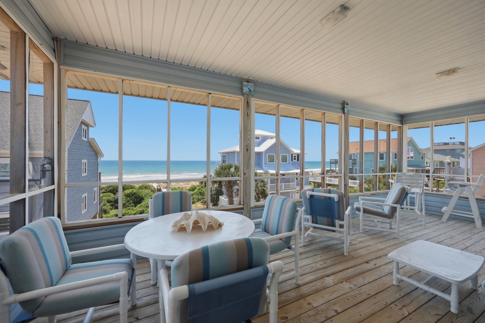 A covered porch with classic beachfront seating invites relaxation. Neighboring homes and coastal views frame the surrounding beach community.