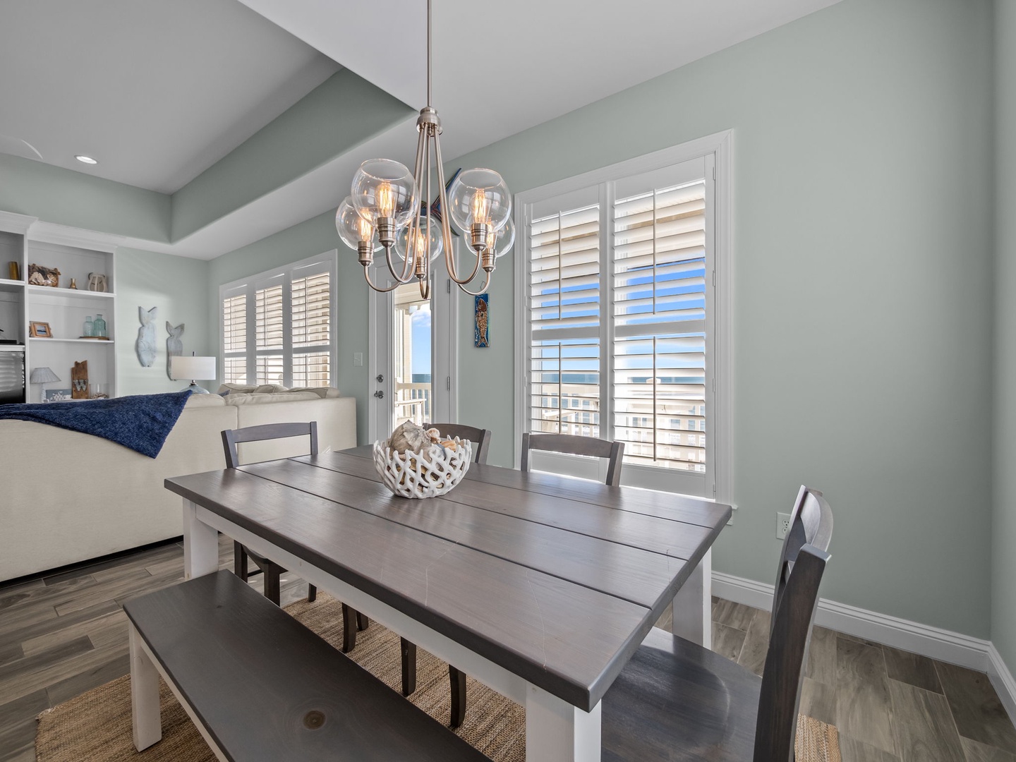 Gather around your elegant dining table where coastal breezes flow through plantation shutters and warm Edison bulb lighting creates perfect dinner ambiance.