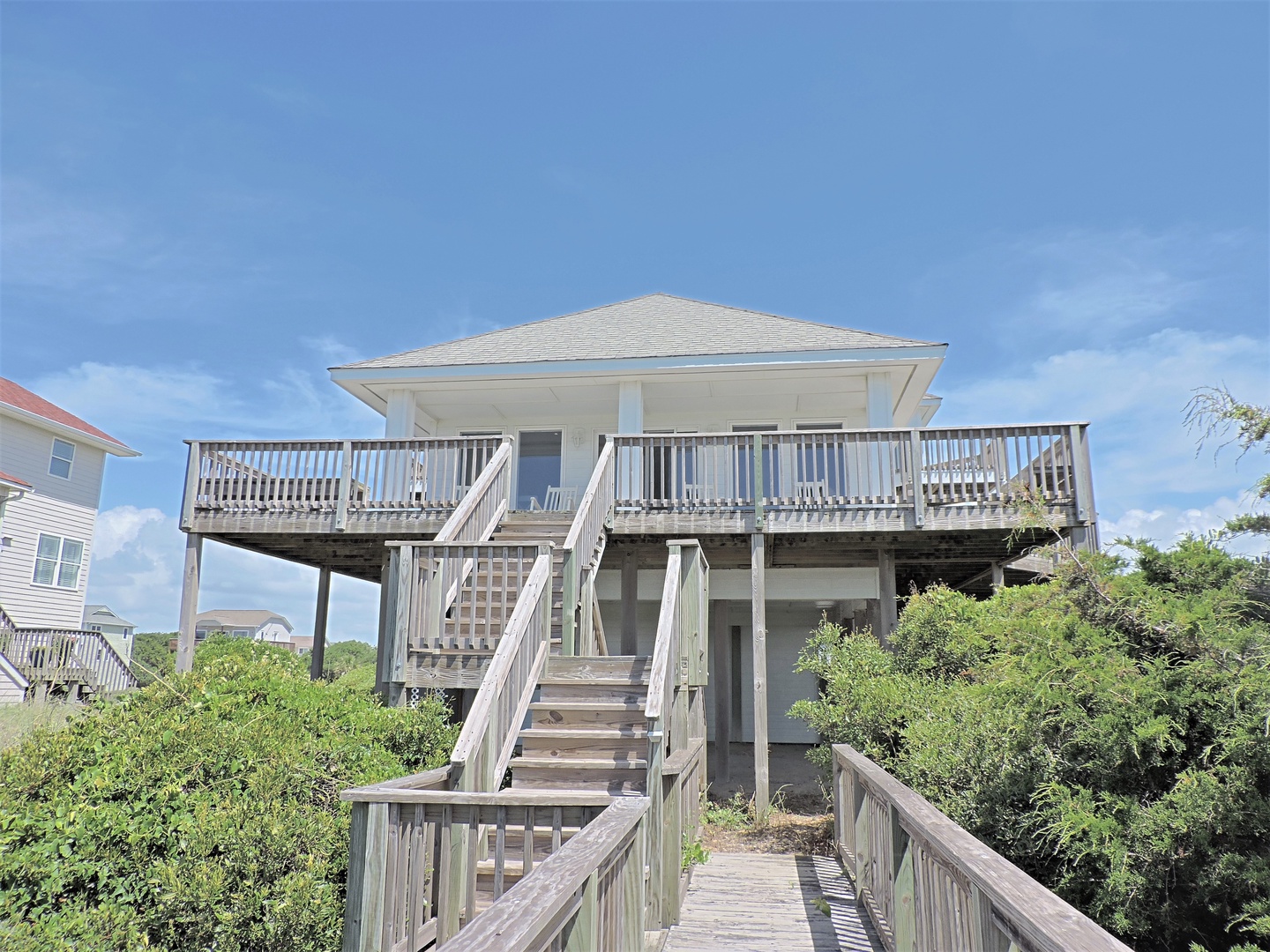 Elevated beach house with wraparound deck and wooden walkway, nestled among coastal vegetation under clear blue skies.
