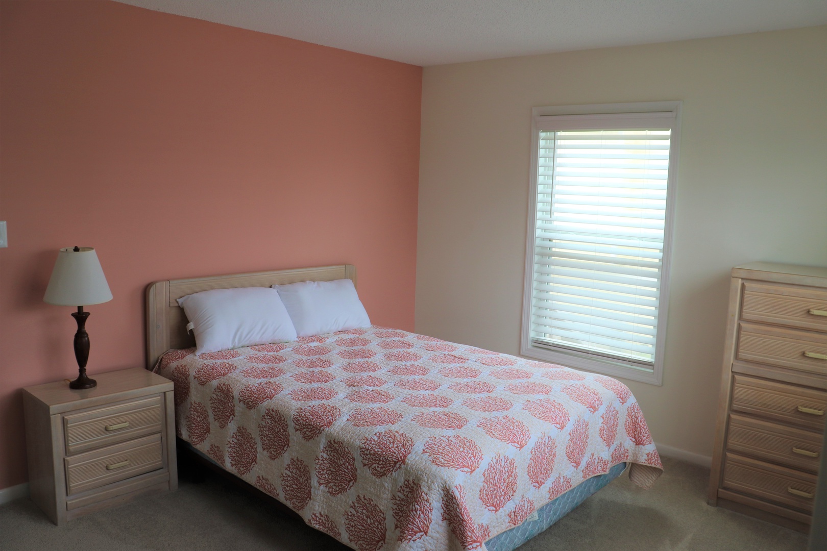 Rest peacefully in your cozy bedroom featuring warm coral accents, comfortable furnishings, and natural light streaming through window blinds.