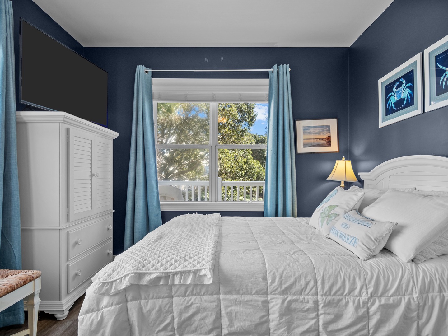 Sink into crisp white linens in your coastal blue bedroom, where morning light filters through windows revealing leafy views.