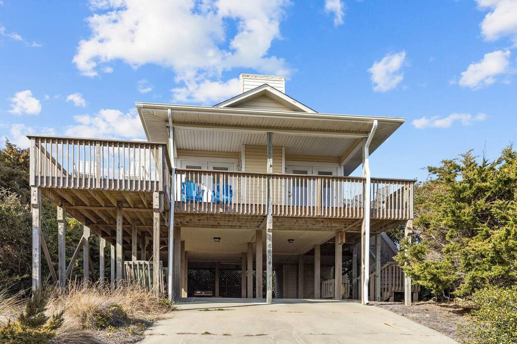 Elevated beach house with spacious wraparound deck and covered parking beneath, surrounded by natural coastal vegetation.