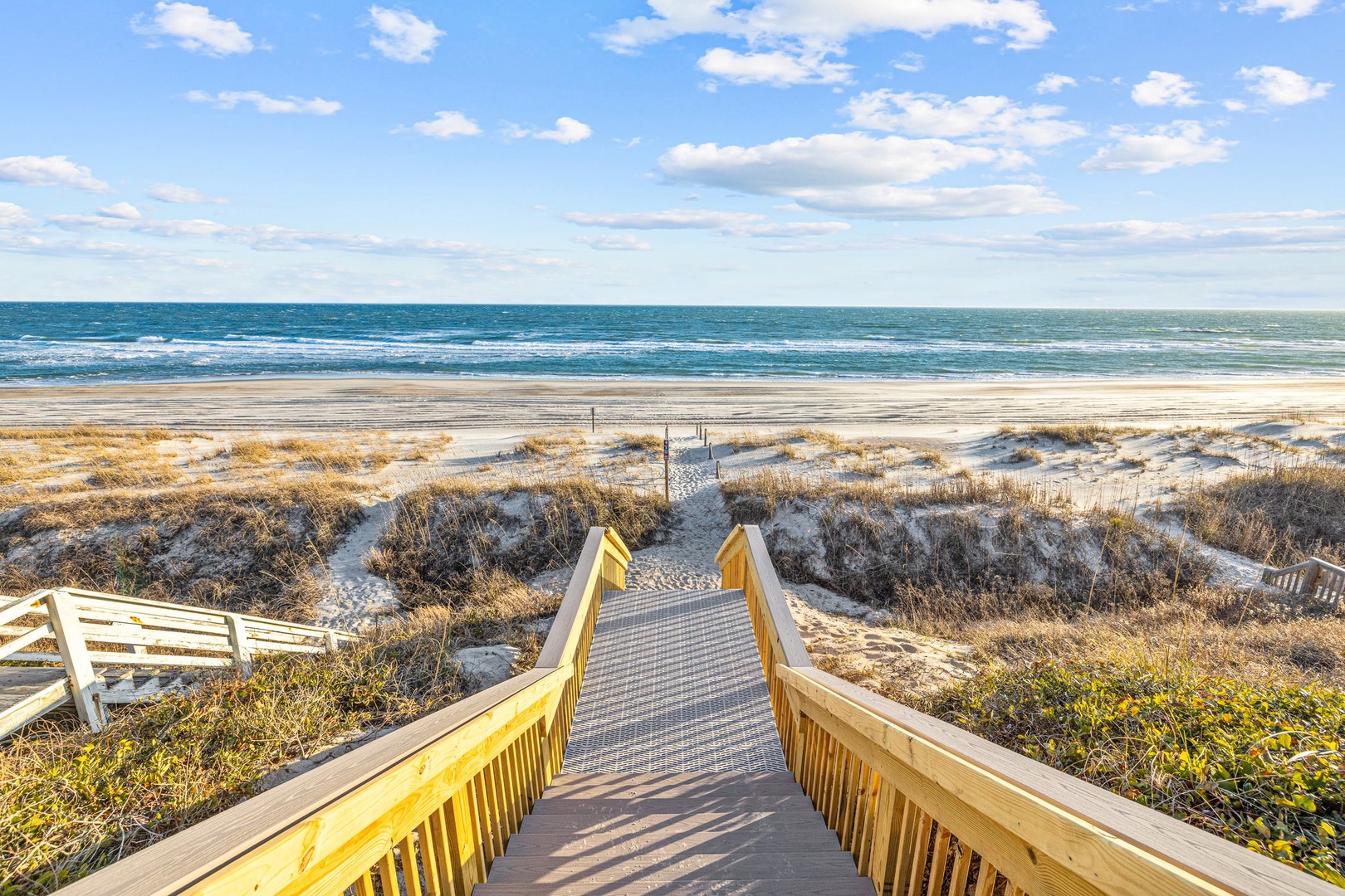 Wooden boardwalk leads through coastal dunes to pristine sandy beach and rolling ocean waves under expansive blue sky.