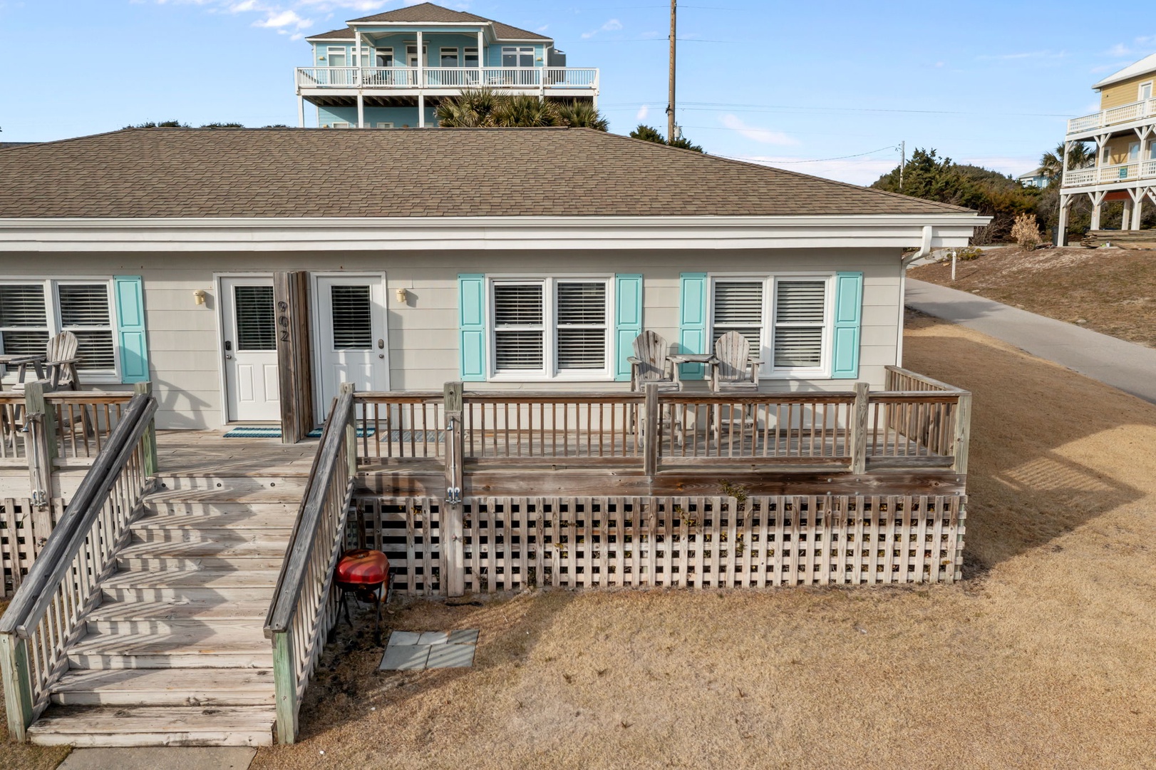 Charming beach cottage with turquoise shutters and expansive deck featuring outdoor seating area in coastal neighborhood setting.