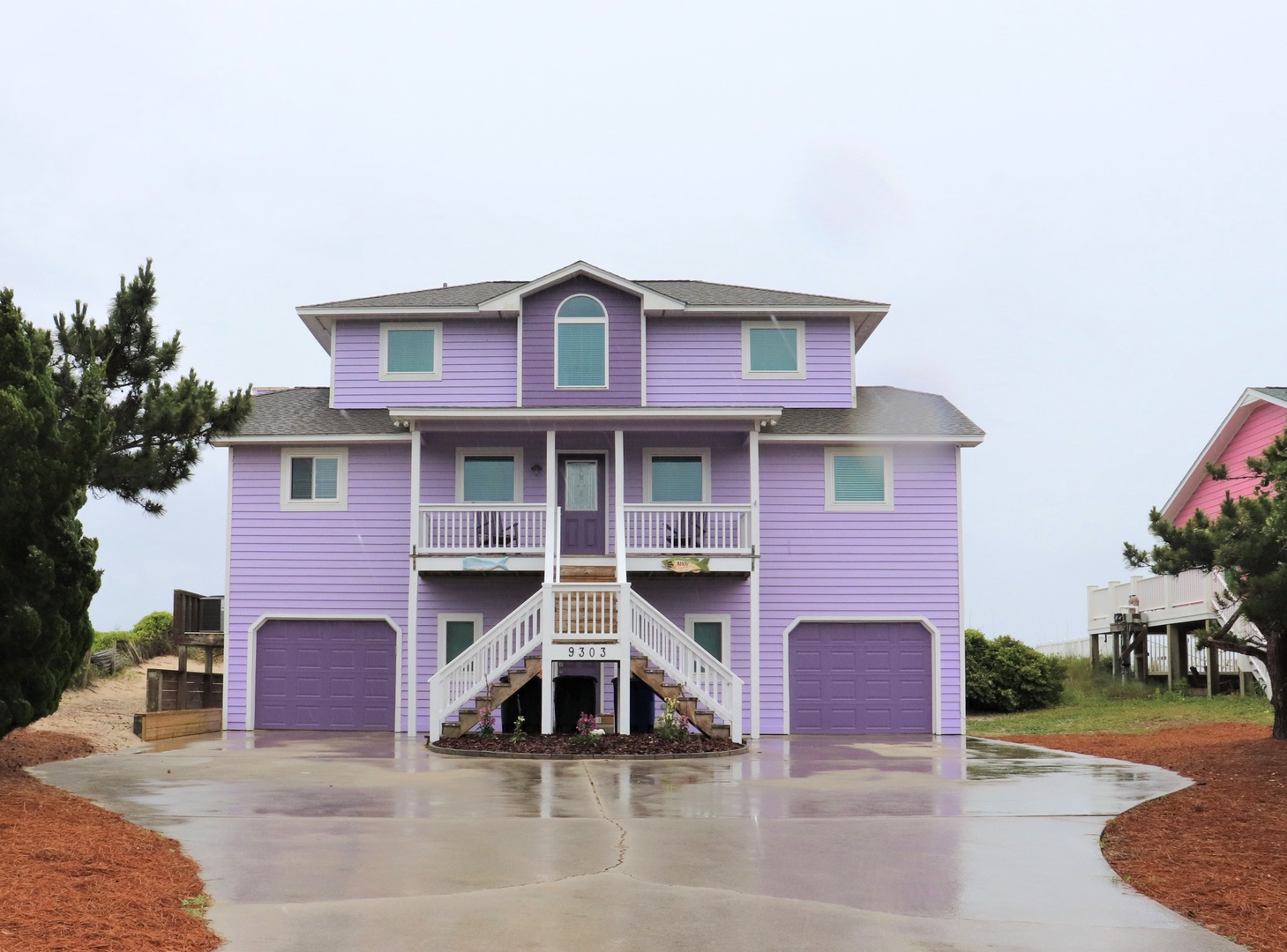 Purple coastal beach house with two-car garage and covered entrance stairs surrounded by landscaped grounds.
