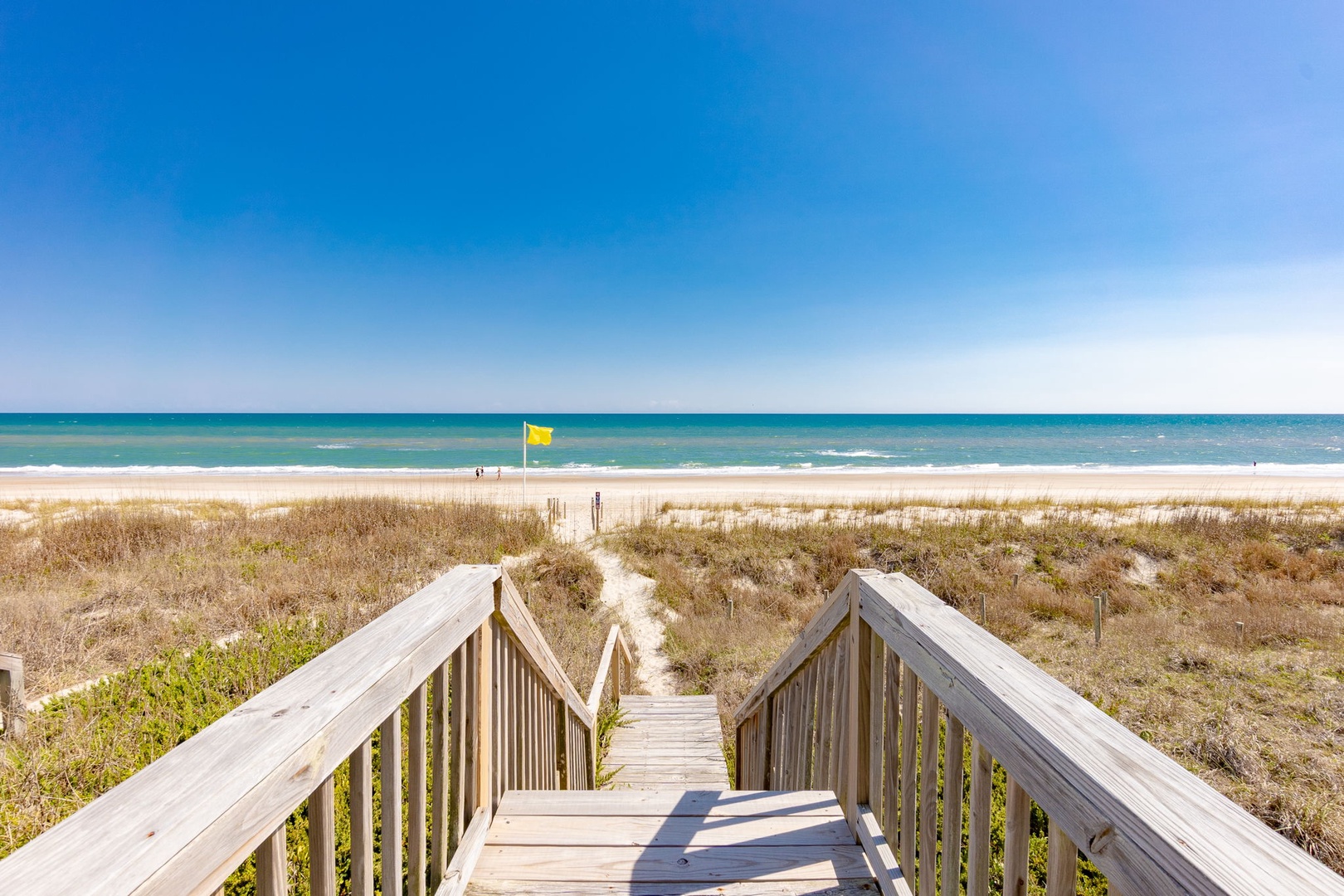 A peaceful boardwalk leads across natural dunes to pristine sandy shores and crystal-clear waters under brilliant blue skies.