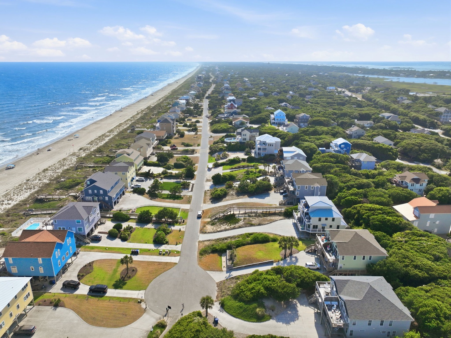Aerial view of a coastal community nestled between ocean and sound, showcasing beachfront vacation homes amid lush greenery and pristine sandy shores.