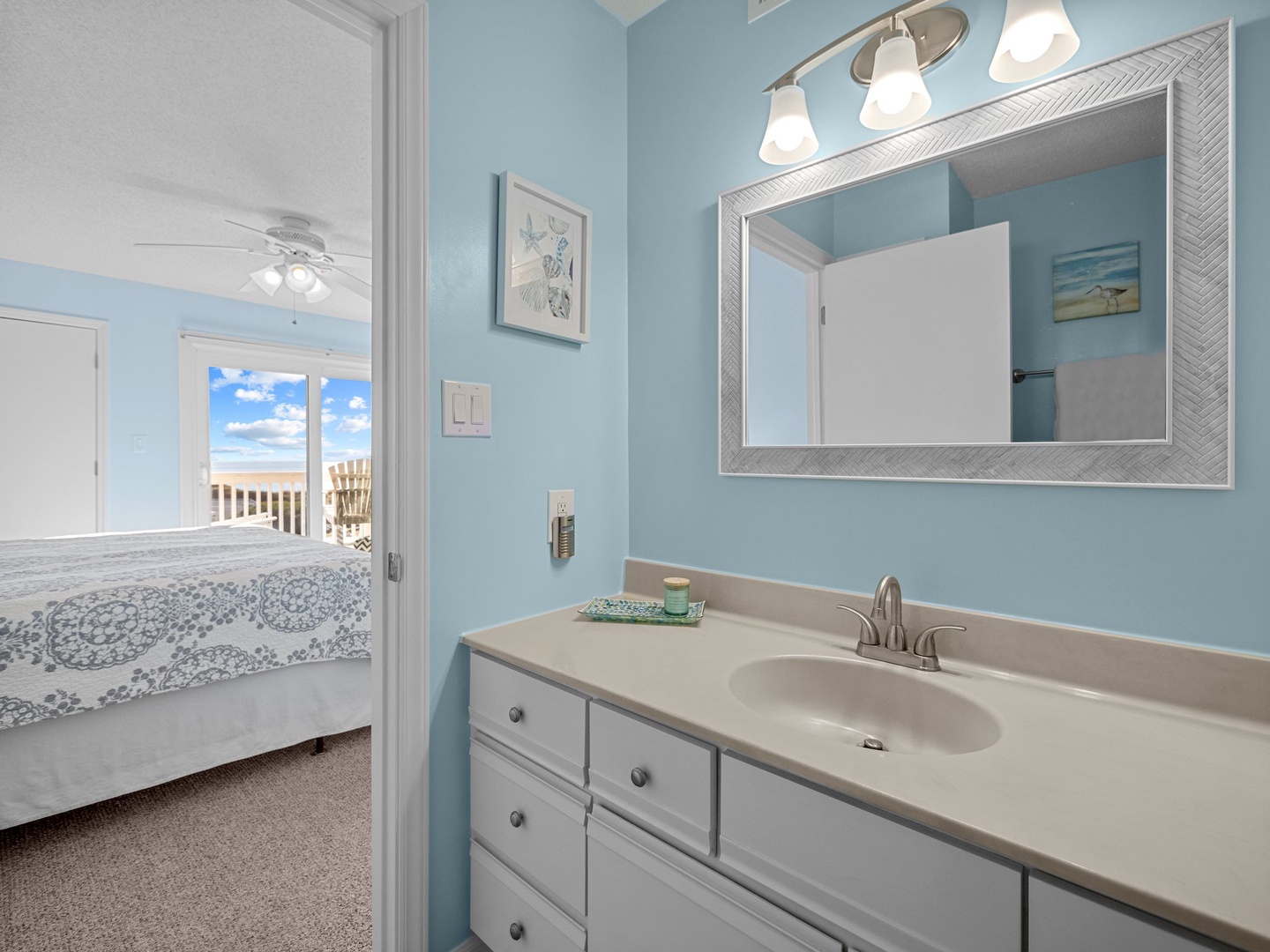 Freshen up in your coastal-themed bathroom featuring soothing blue walls, bright vanity lighting, and ocean views through the bedroom.