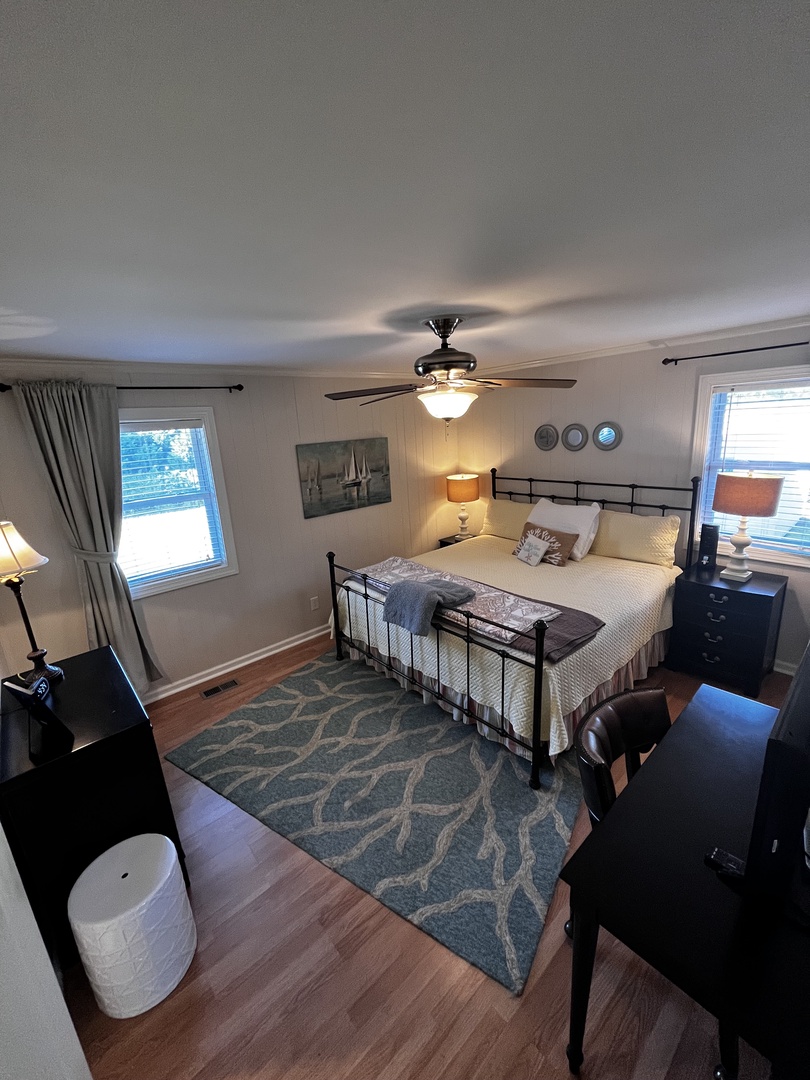 Your bright bedroom features a cozy bed with ocean-themed bedding and natural light streaming through windows.