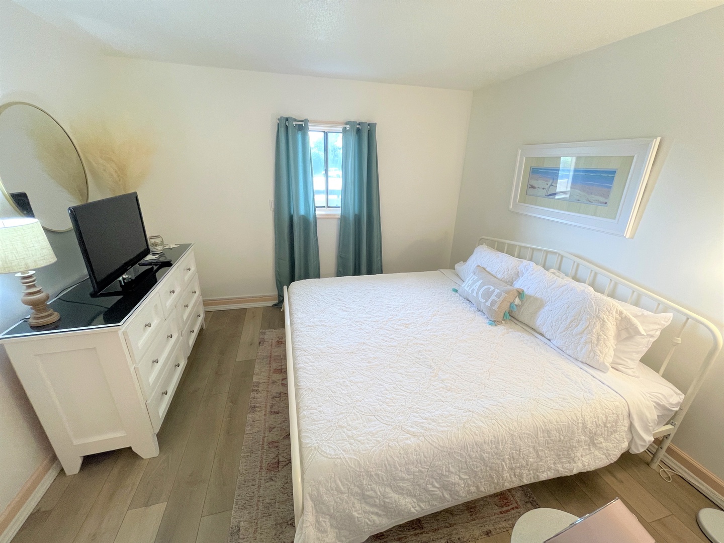 Your serene bedroom retreat features coastal charm with crisp white linens, a flatscreen TV for relaxation, and peaceful teal curtains filtering gentle light.