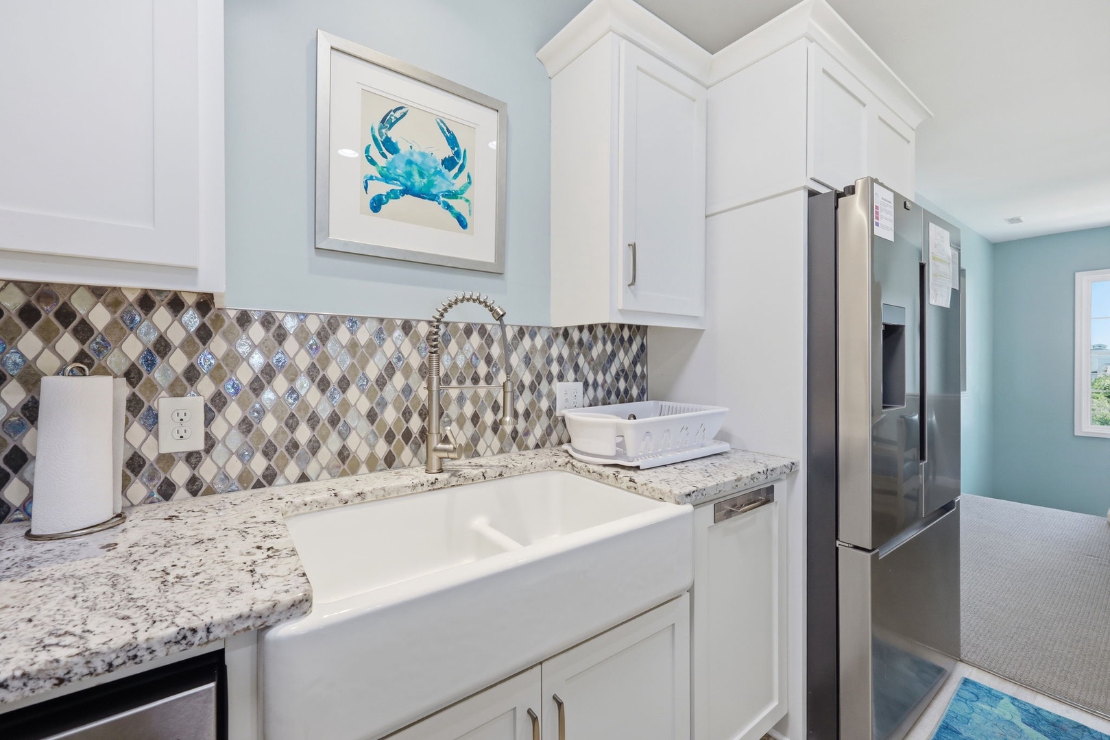 A charming coastal-inspired kitchen where you'll love prepping meals, featuring granite counters, custom backsplash, modern appliances, and cheerful blue crab artwork above the farmhouse sink.