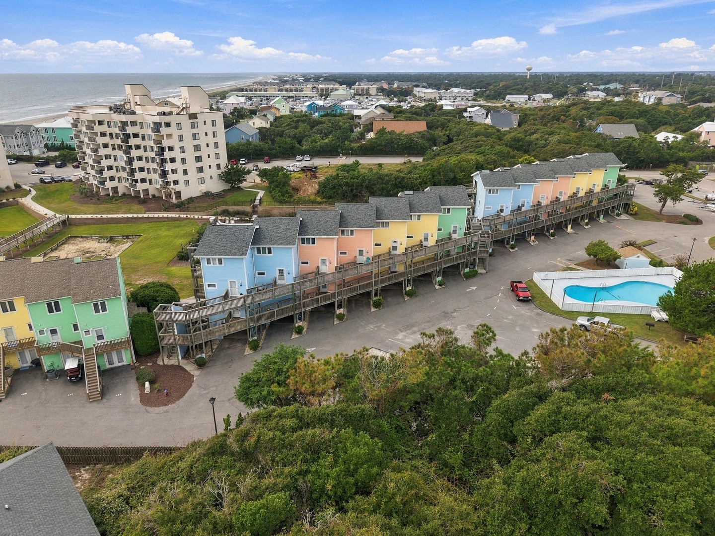 Aerial view of a colorful beachside community featuring multi-story buildings, a swimming pool, and parking areas. The property sits in a coastal neighborhood with tree-lined streets and ocean views in the distance.