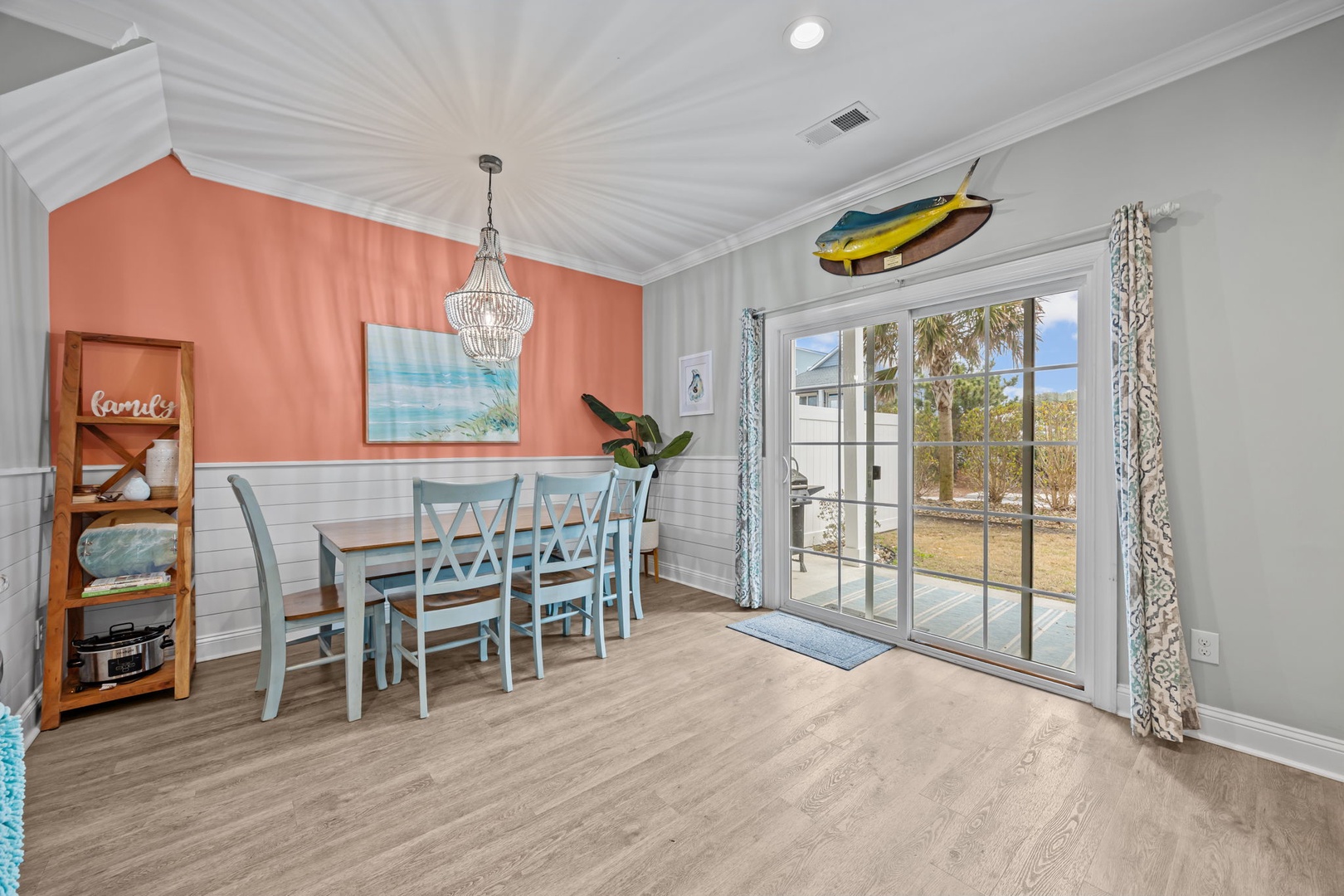 Your bright dining space features coastal charm with turquoise chairs and beach artwork, opening to peaceful outdoor views through sliding glass doors.