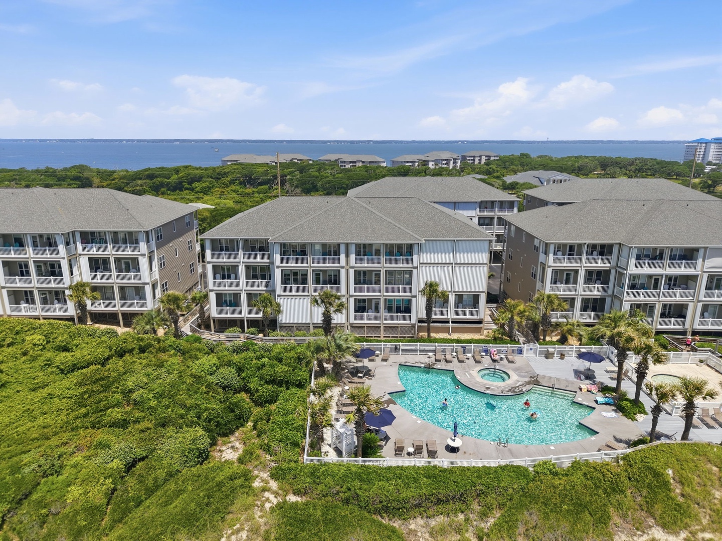 A coastal resort community featuring multi-story residential buildings, a central pool with hot tub, and lush green surroundings with ocean views. The property offers convenient beach access through natural dune vegetation.