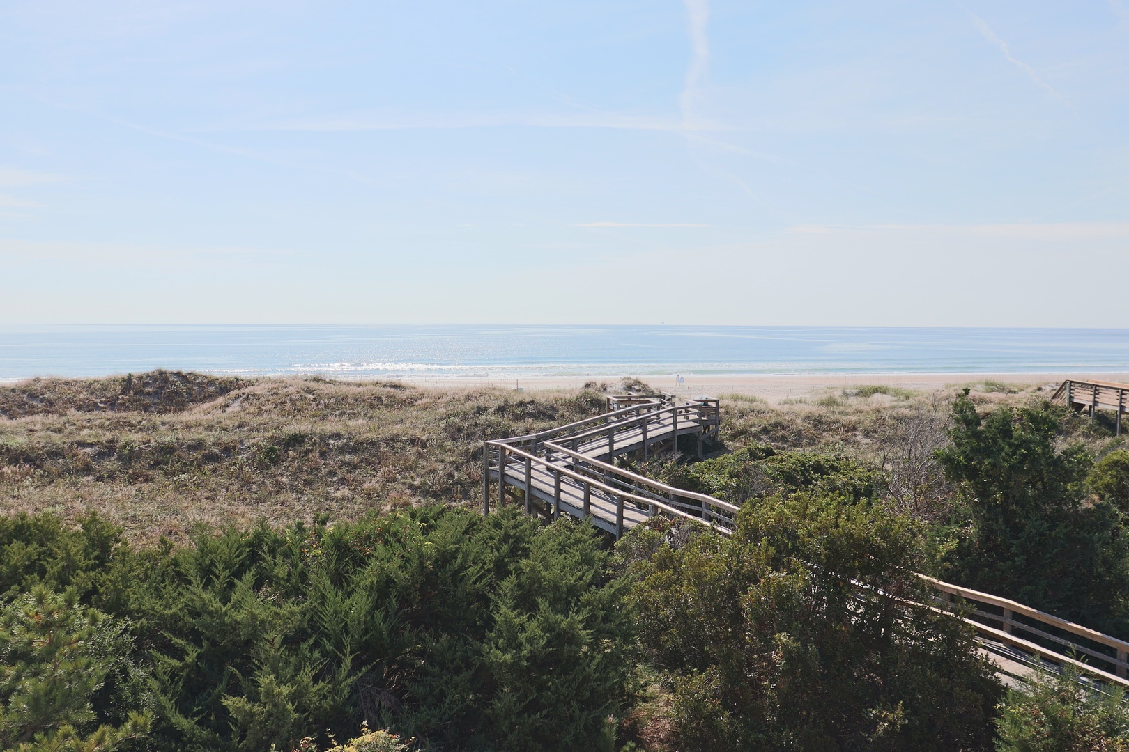 Wooden boardwalk winds through coastal dunes toward pristine sandy beach and sparkling ocean waters under clear blue skies.