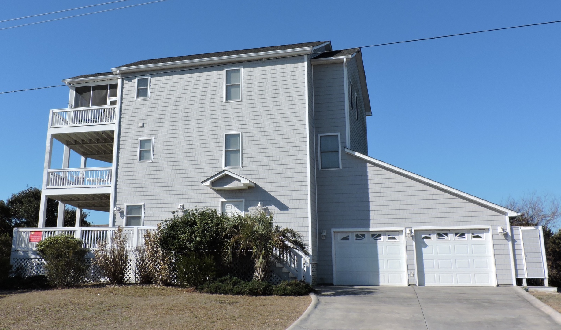 A three-story coastal home with spacious balconies and garage parking, surrounded by mature landscaping under clear blue skies.
