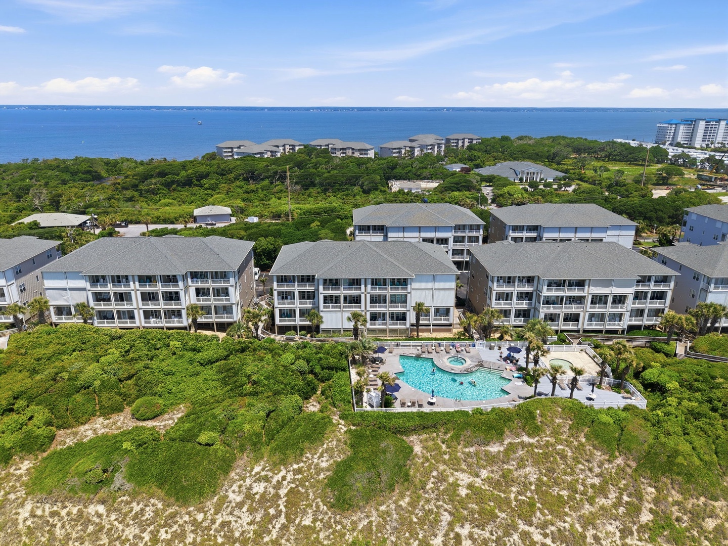Aerial view of a coastal vacation resort featuring multiple modern buildings surrounded by lush greenery, with a central pool area and the ocean visible beyond.
