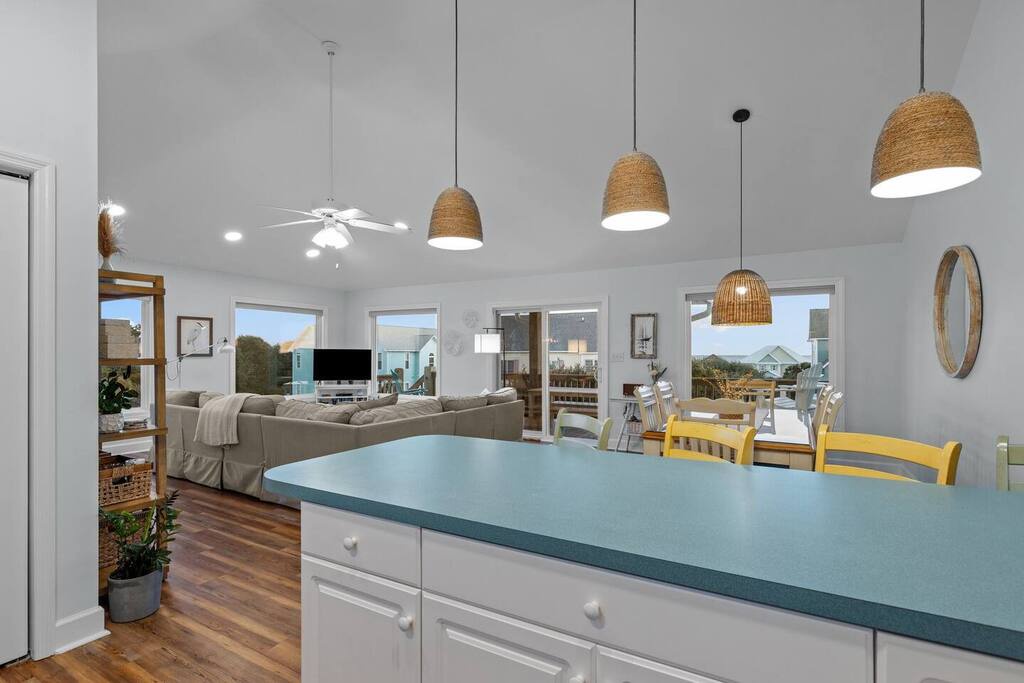 Your bright open-plan kitchen flows seamlessly into the living area, with natural woven pendant lights creating warmth overhead and colorful dining chairs adding charm.
