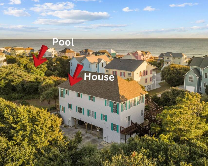 Elevated coastal home with pool, nestled among beach houses with ocean backdrop and lush vegetation.