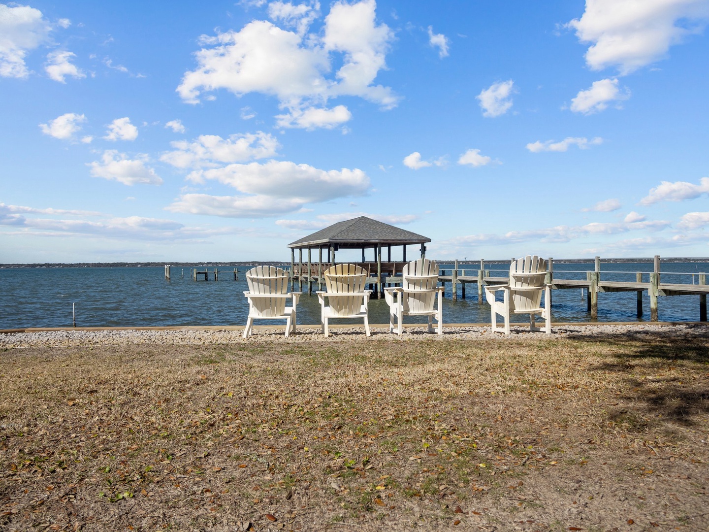 Waterfront setting with Adirondack chairs facing a peaceful lake, private dock, and covered gazebo under open blue skies.