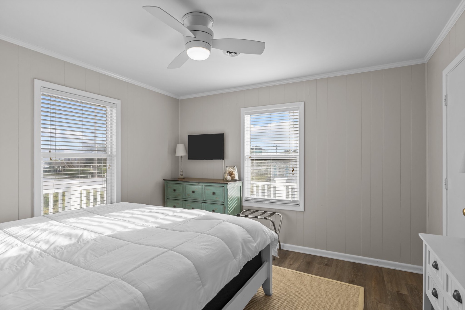 Wake up refreshed in this bright coastal bedroom with soft linens, gentle ceiling fan breeze, and peaceful neighborhood views through dual windows.