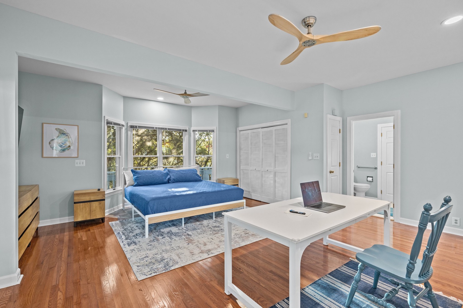 Relax in your serene bedroom with calming blue tones, ceiling fans, and a dedicated workspace. Natural light streams through windows.