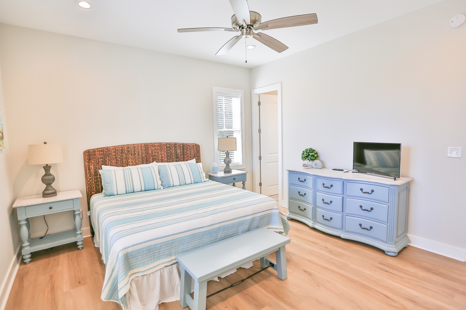 Relax in your coastal-inspired bedroom with crisp blue linens, wicker headboard, and gentle ceiling fan creating the perfect sea breeze atmosphere.