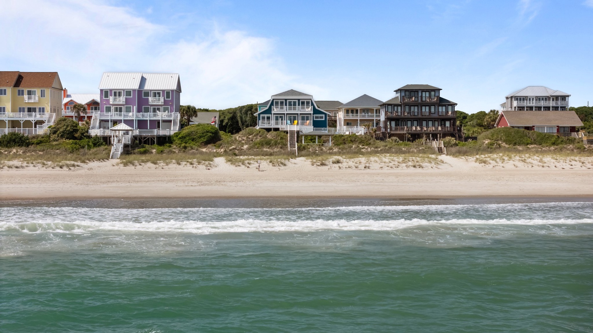 Charming beachfront homes line the sandy shore with gentle waves rolling in under bright blue skies.
