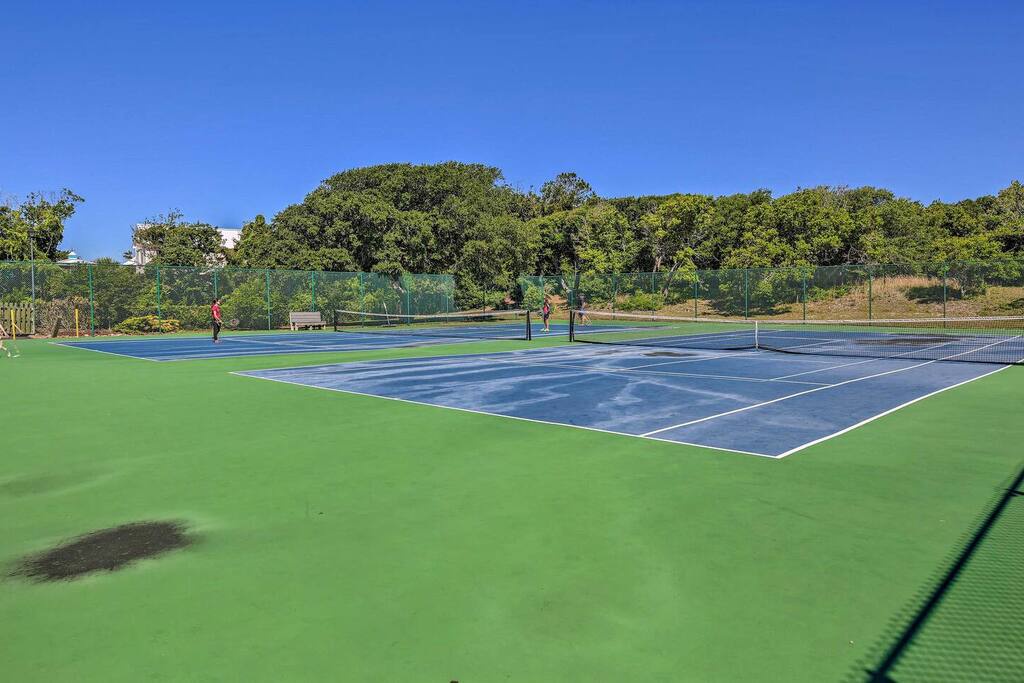 Professional tennis courts with pristine blue surfaces and green surrounds, set against mature trees and clear skies.