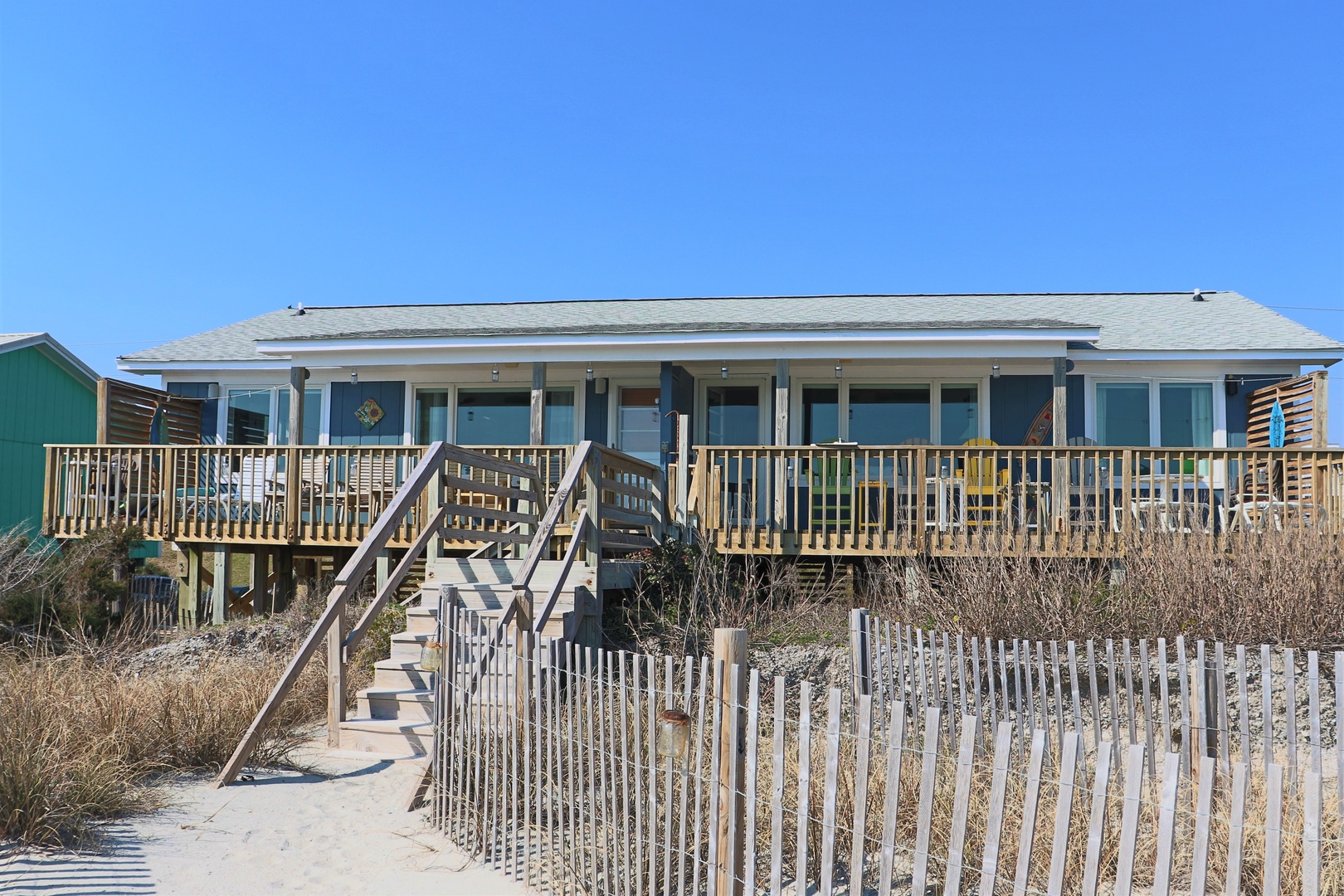 Beachfront cottage with elevated deck and stairs leading from sandy dunes under clear blue skies.