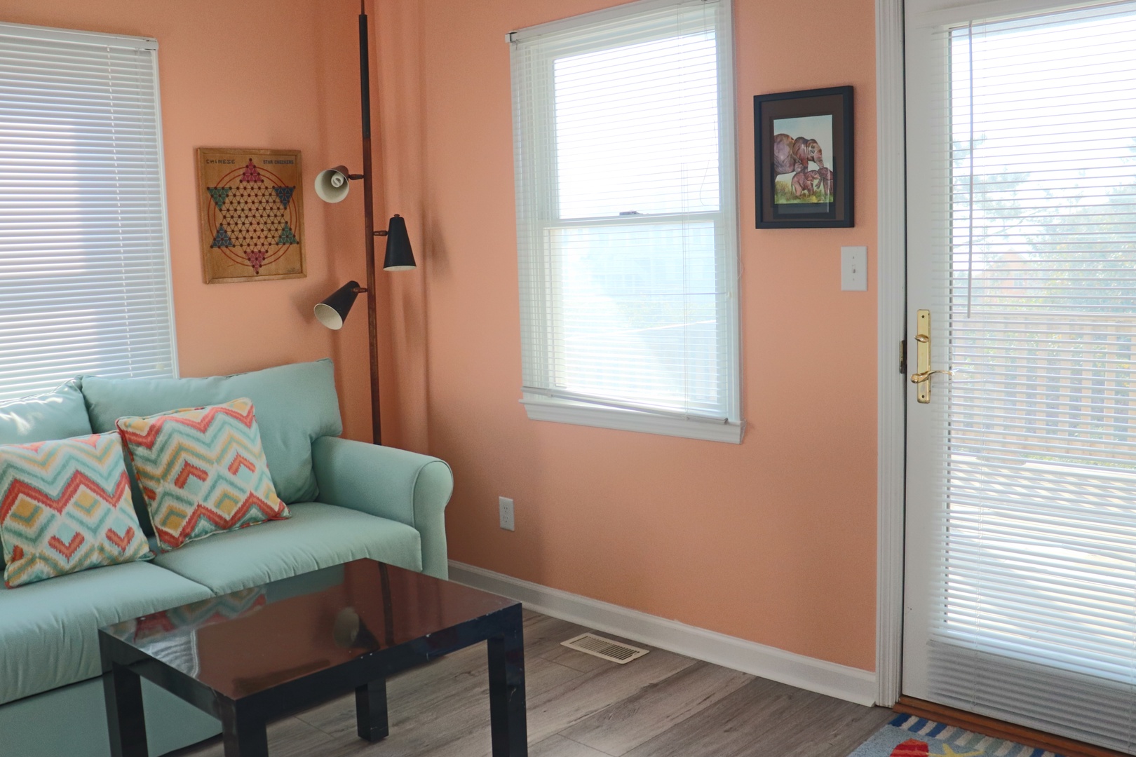 Your bright living space features cheerful coral walls, comfy mint sofa with colorful pillows, and large windows flooding the room with natural light.