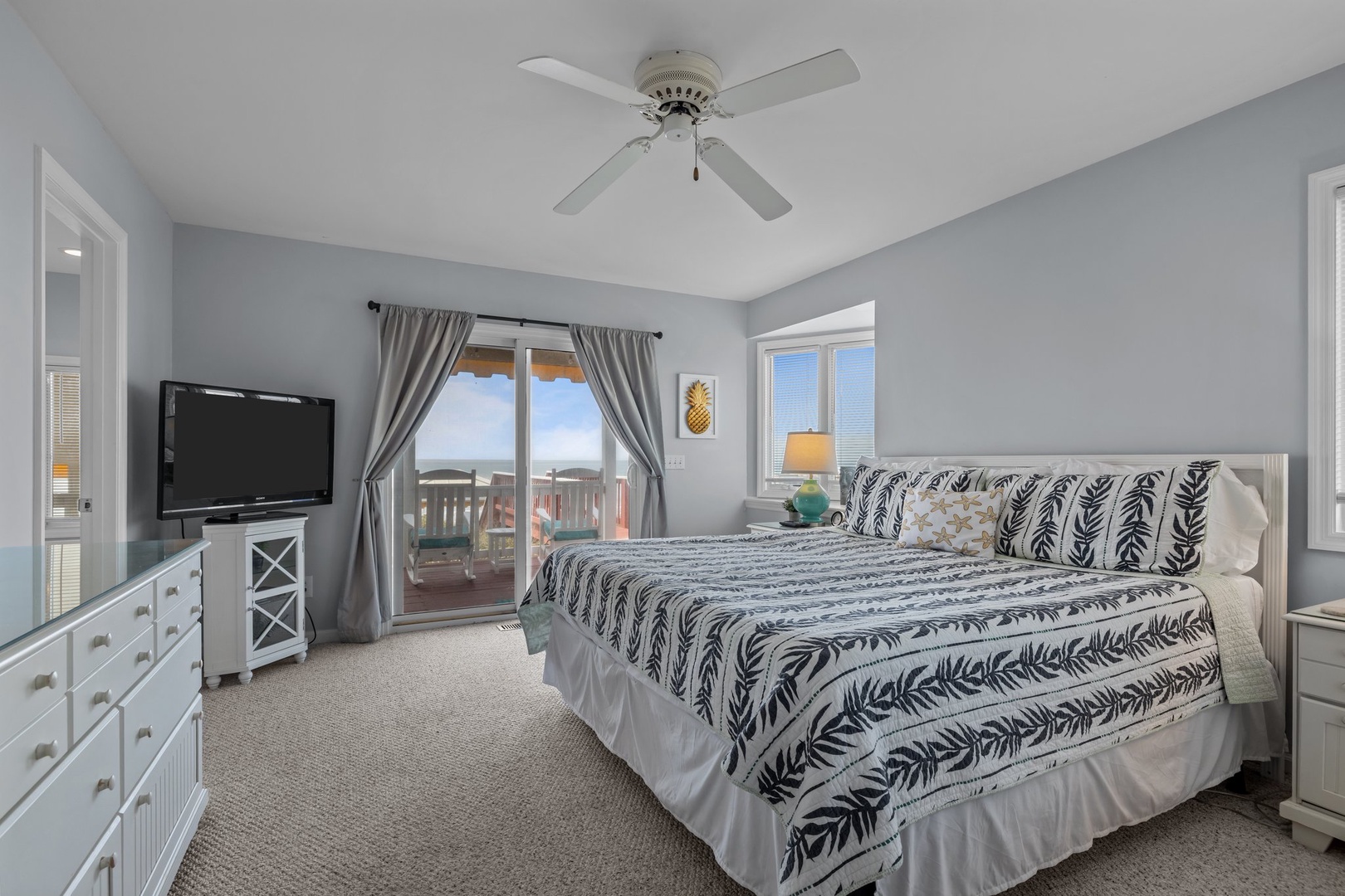 Wake up in your serene coastal bedroom with calming blue-gray walls, ceiling fan comfort, and a private balcony overlooking the beach.