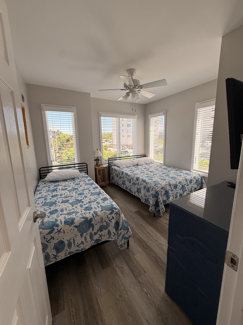 Bright natural light floods your bedroom through skylights and large windows, creating an airy retreat with charming blue-toned beds and cozy wood floors.