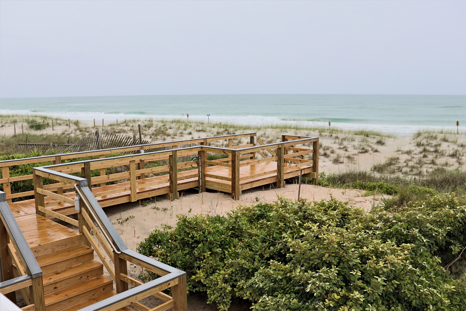 Wooden boardwalk leads through dunes to pristine beach with gentle waves and distant figures along the shore.