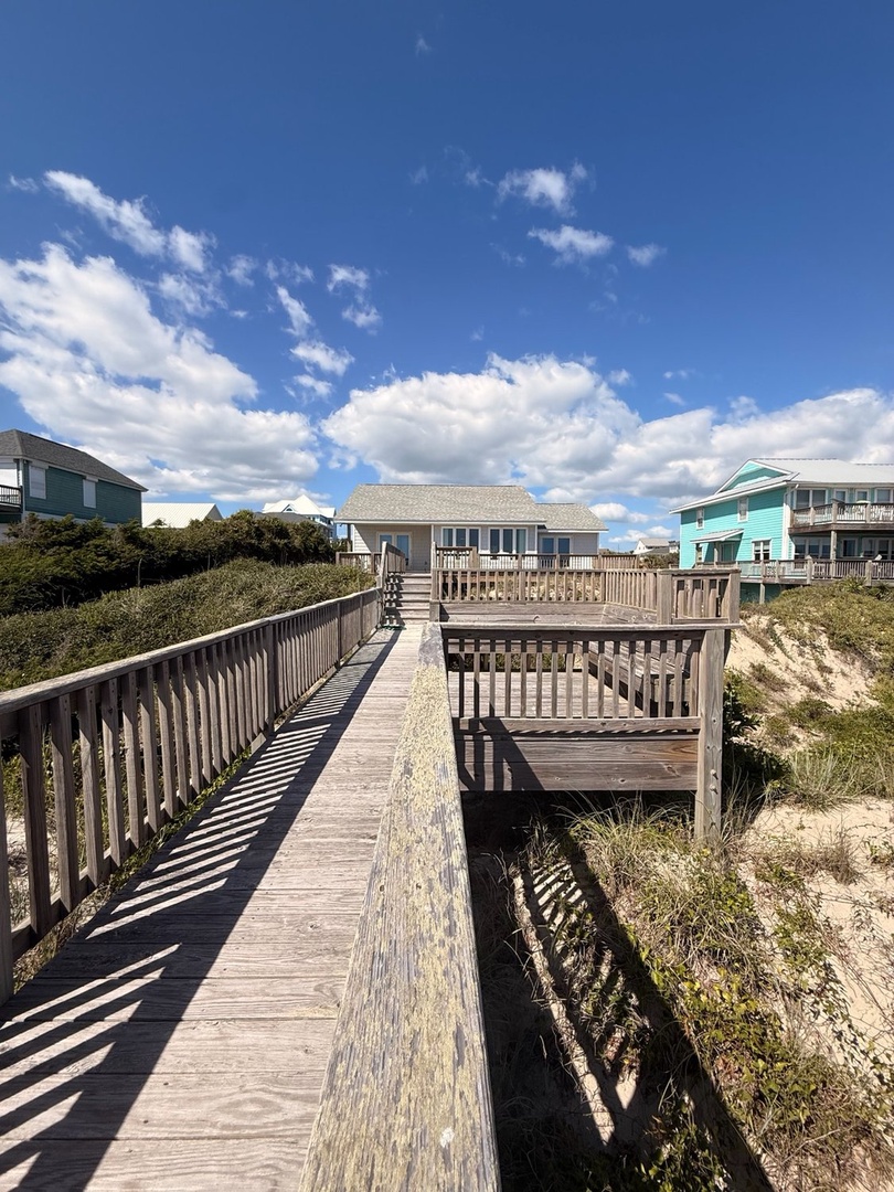 A private boardwalk leads from the property directly through coastal dunes to nearby beach access, with neighboring homes and clear blue skies completing the serene coastal setting.