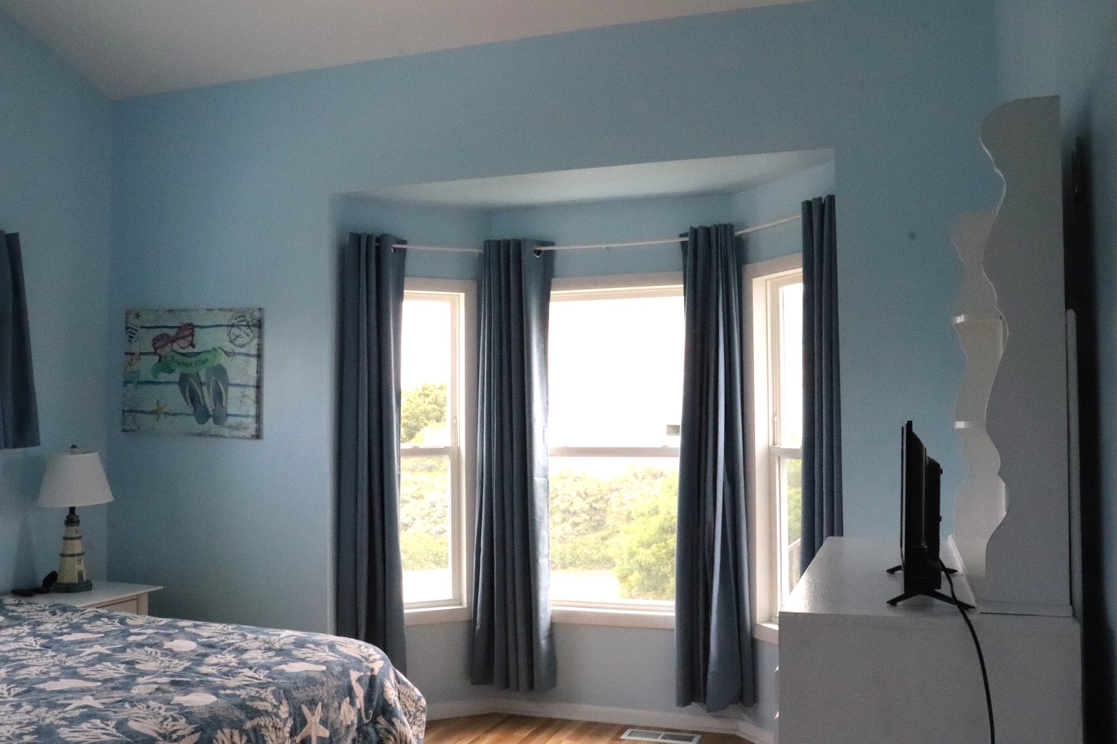 Unwind in your tranquil bedroom retreat with soothing blue tones, natural light streaming through bay windows, and leafy views inviting peaceful moments of rest.