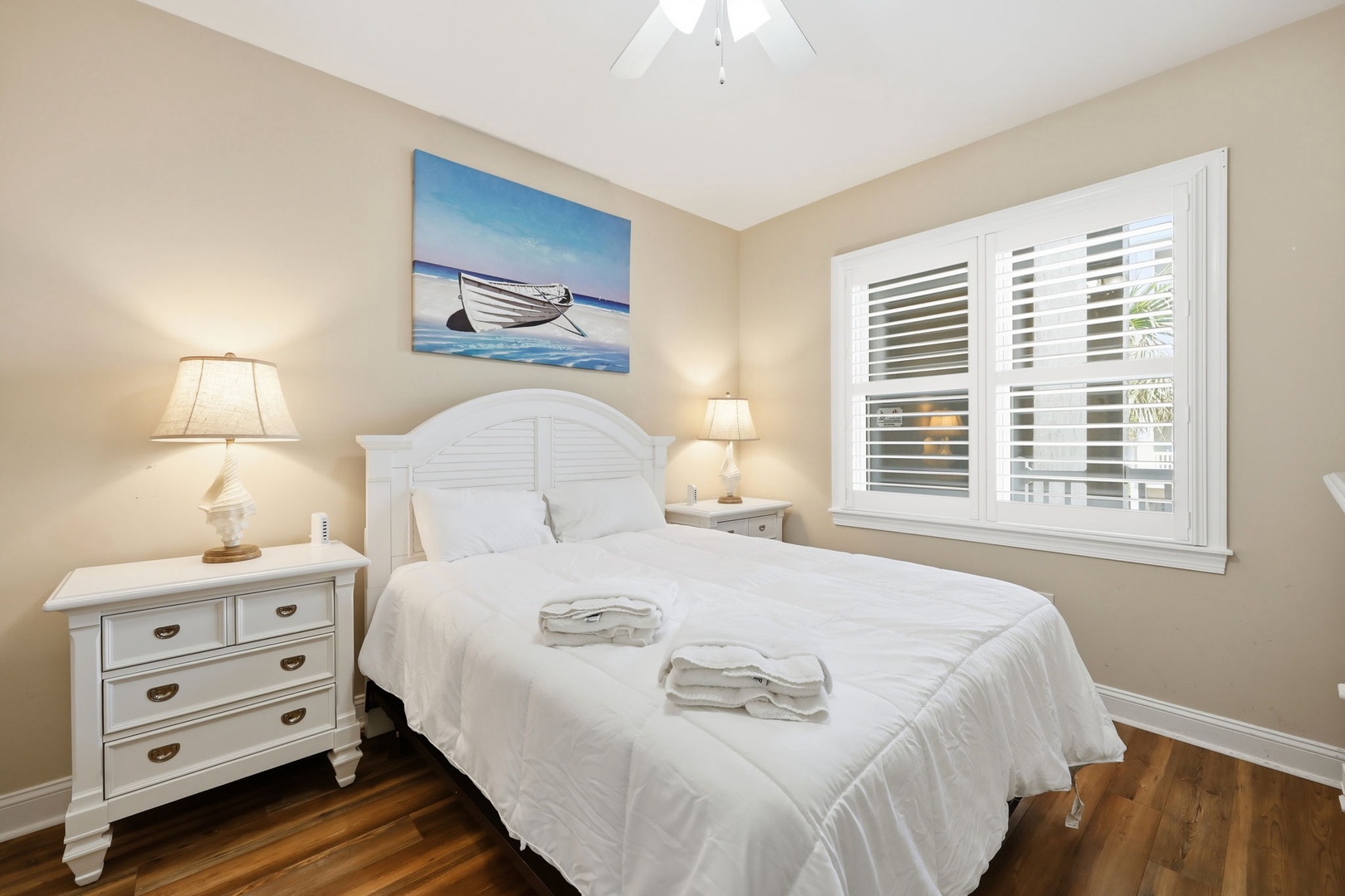 Slip into crisp linens and drift off beneath a coastal boat painting. Your bedroom features a plush bed flanked by nightstands, warm lighting, and soft natural light filtered through plantation shutters.