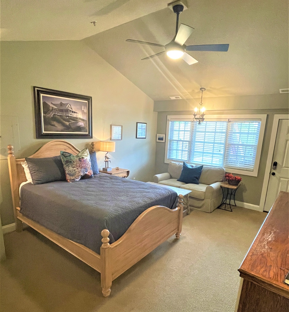 Drift off peacefully in your spacious bedroom featuring a cozy seating area, ceiling fan, and gentle natural light through plantation shutters.