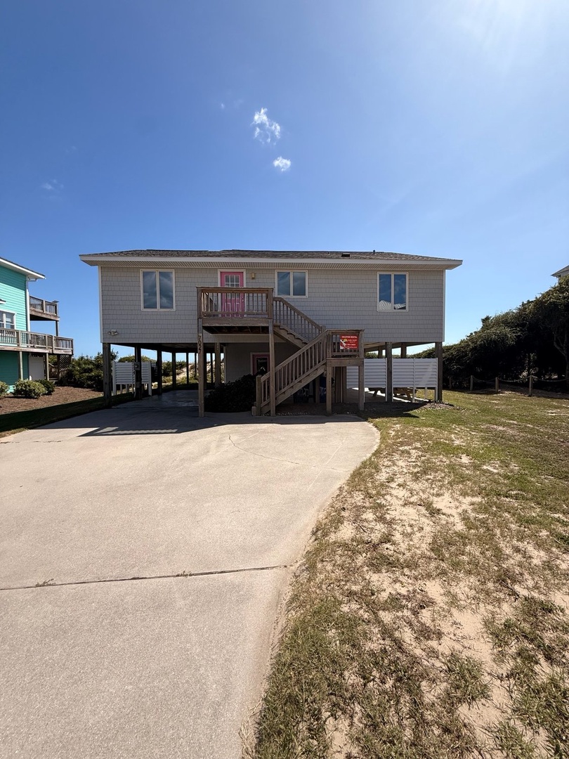 Elevated beach home with ample parking and covered ground-level storage near coastal vegetation.