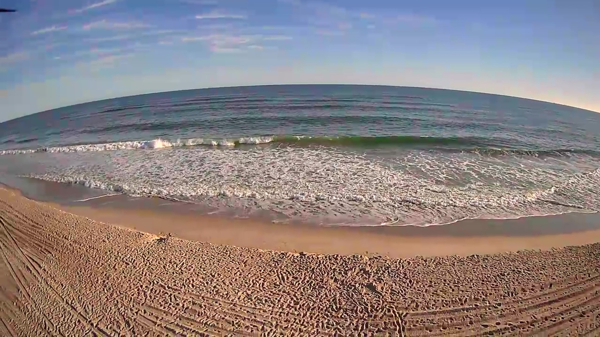 Wide curved beach with gentle waves rolling onto golden sand under a cloudy sky.
