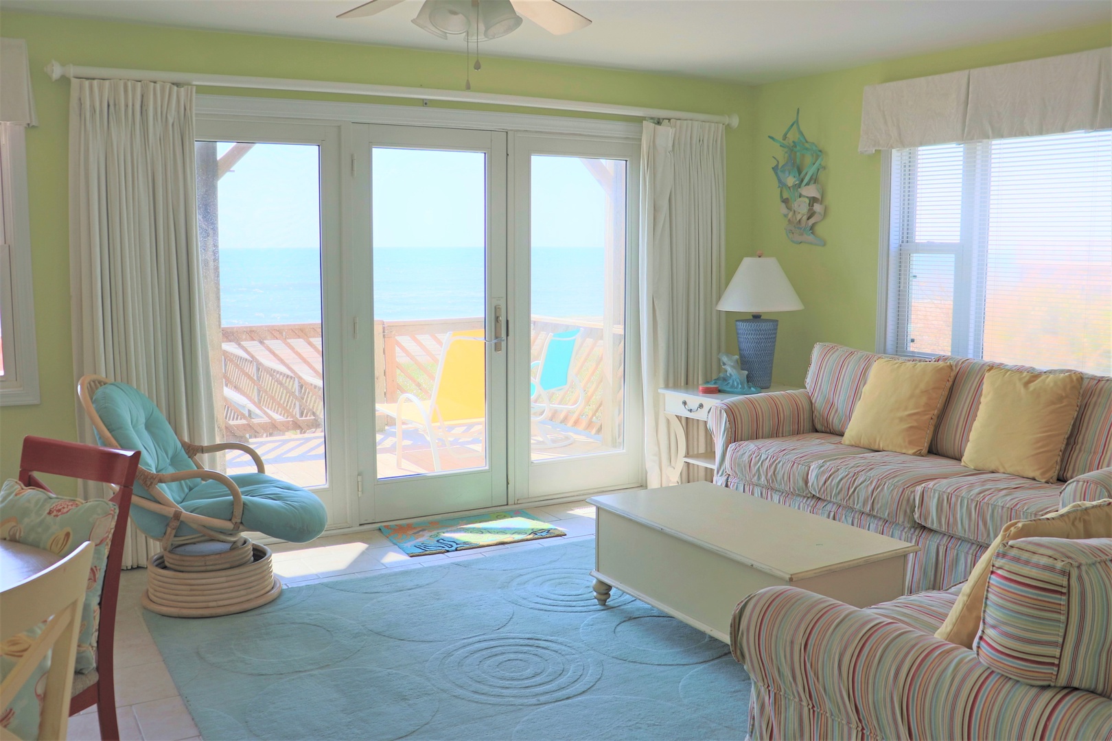 Relax in your bright coastal living room with striped seating, ceiling fan, and stunning sea views through floor-to-ceiling doors.