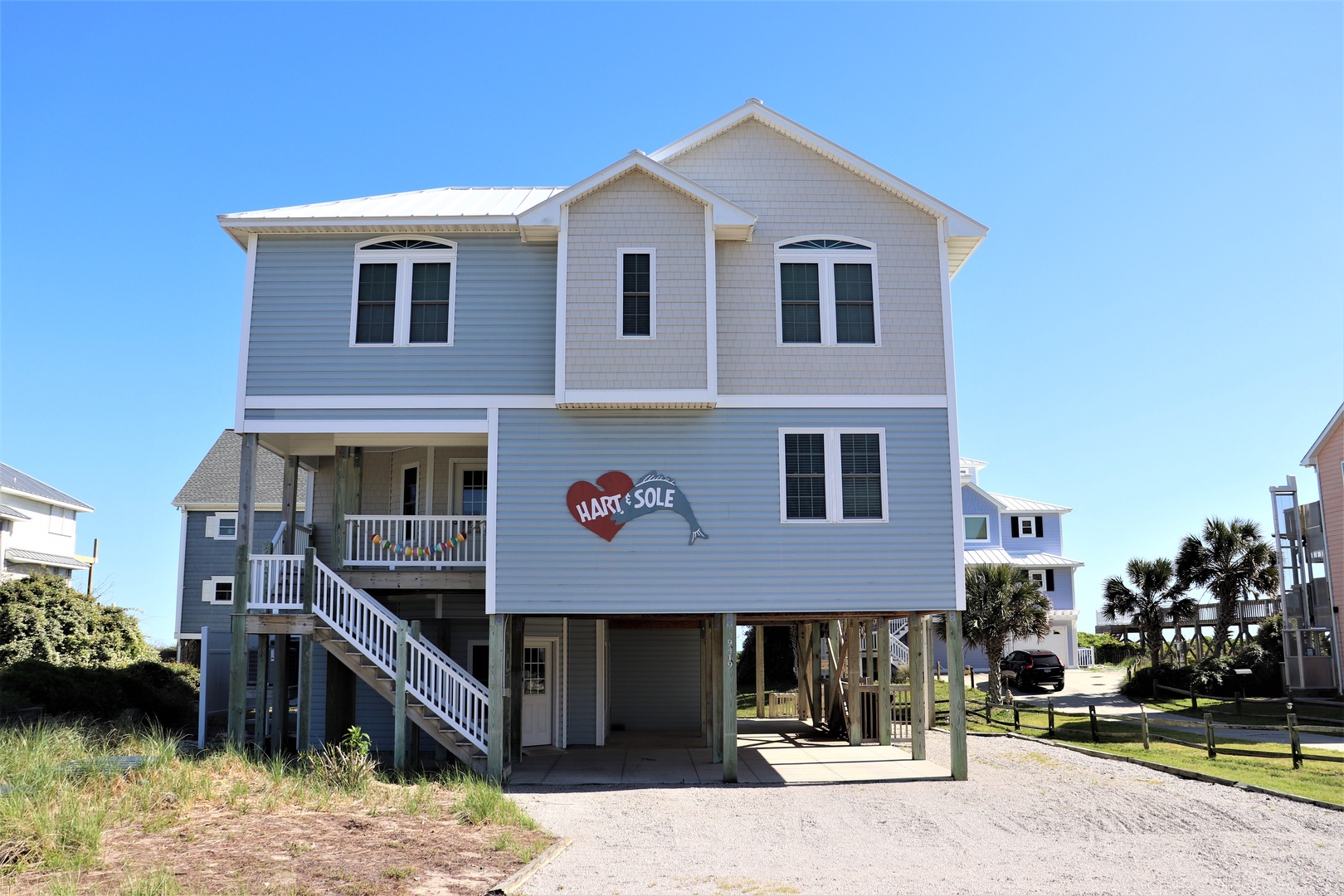 Coastal vacation home featuring raised design and covered parking, situated in a peaceful beachside neighborhood with palm trees and blue skies.
