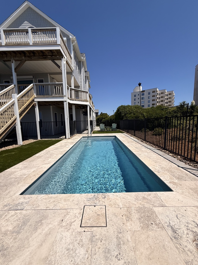 A modern infinity-edge pool nestled against sleek architecture and lush greenery, set on warm stone pavers under clear blue skies—perfect for soaking up the sun in style.