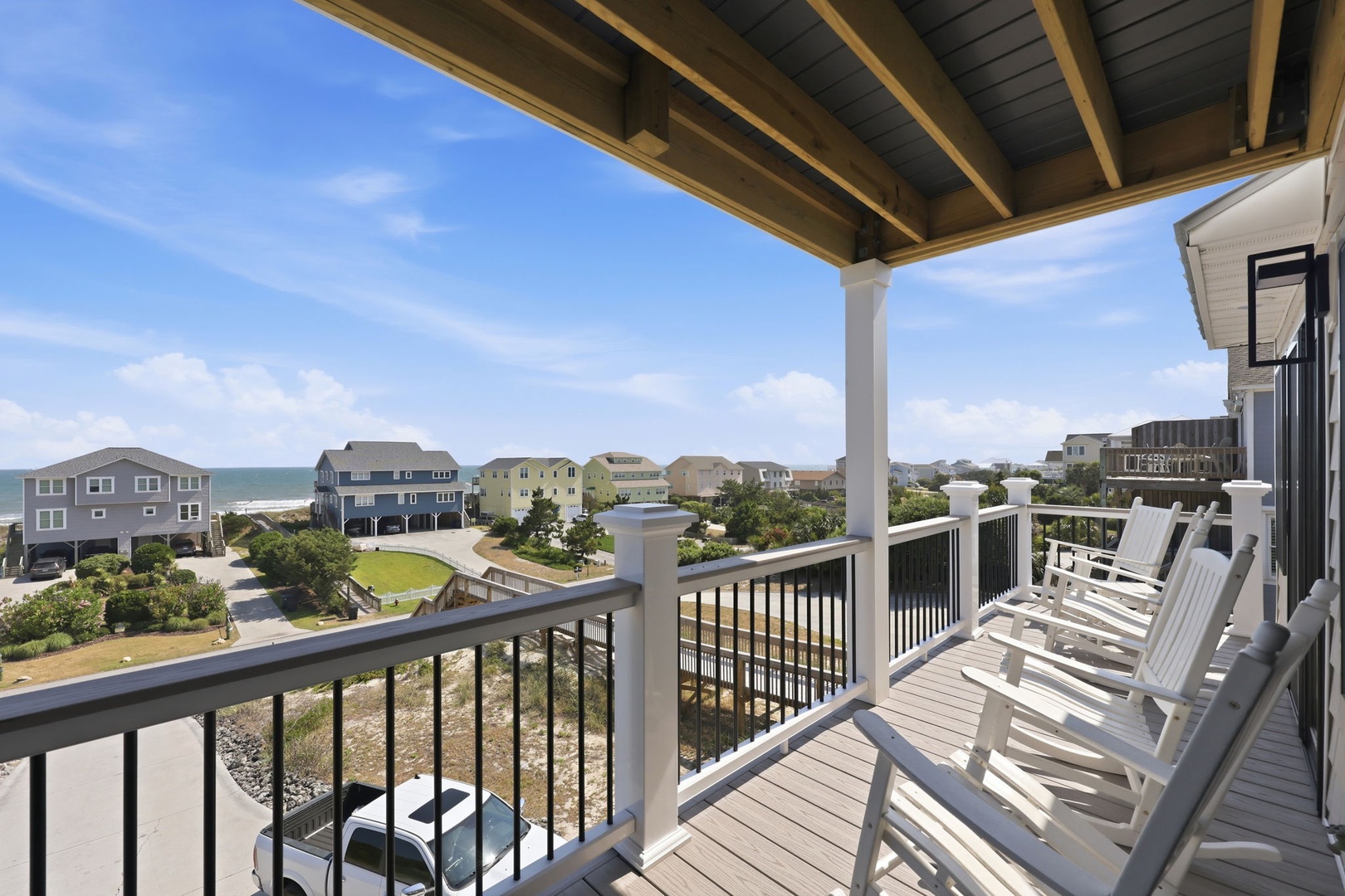 This covered balcony features rocking chairs with coastal views of nearby beach homes, ocean waters, and surrounding landscape—ideal for relaxing mornings and evenings in this seaside setting.