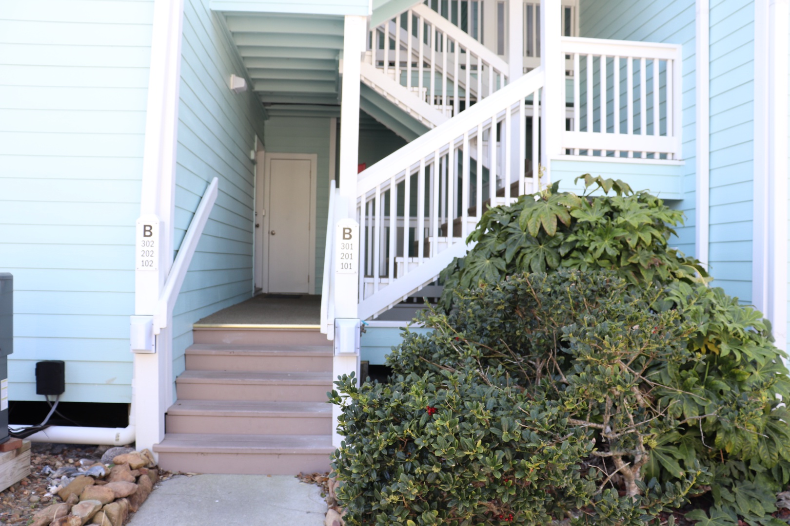 Light blue property building with white railings and landscaped entrance areas featuring unit numbers and outdoor stairs.
