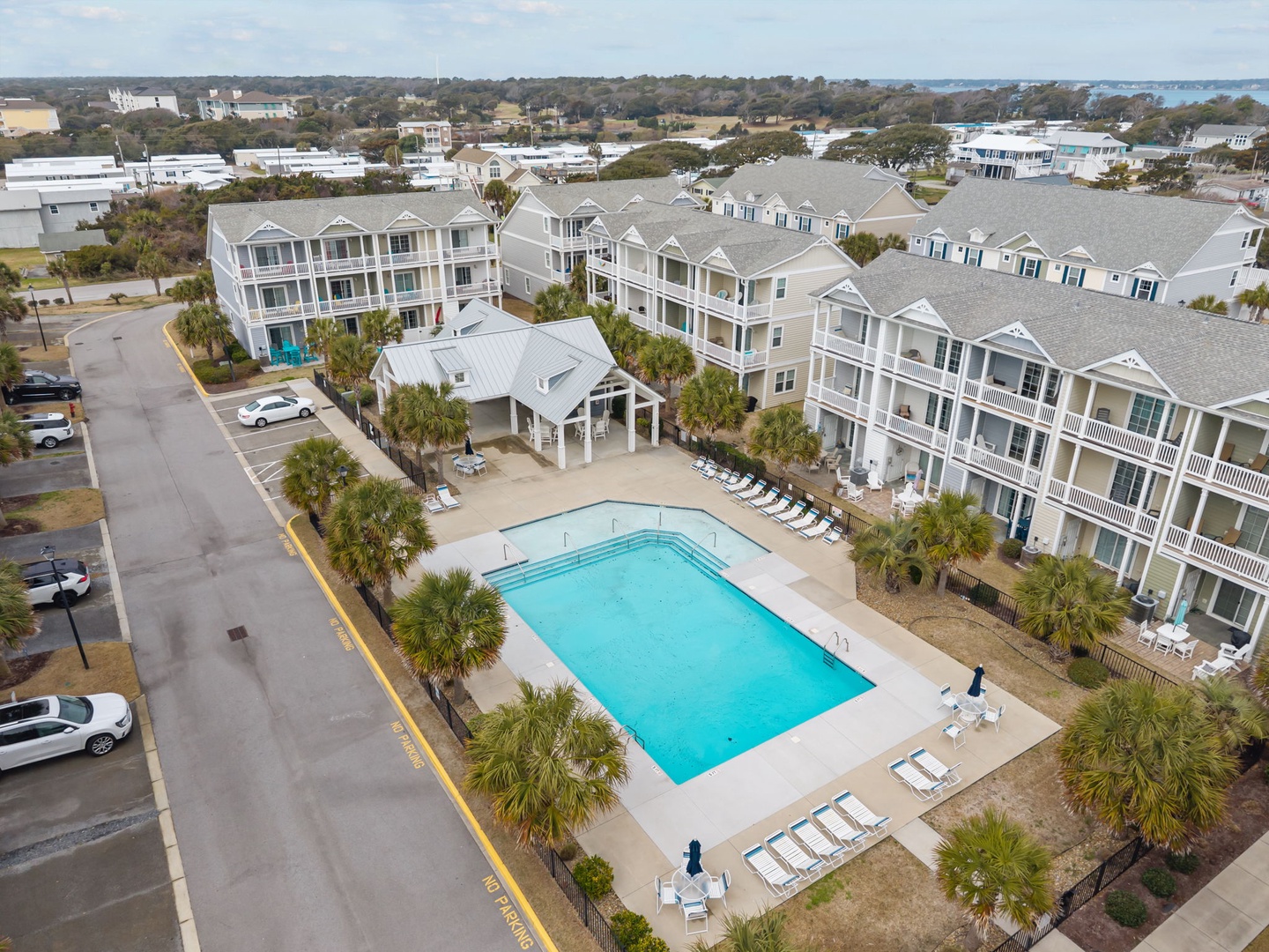 Aerial view of beachside resort complex with swimming pool, multi-story buildings, and palm-lined grounds near coastal community.