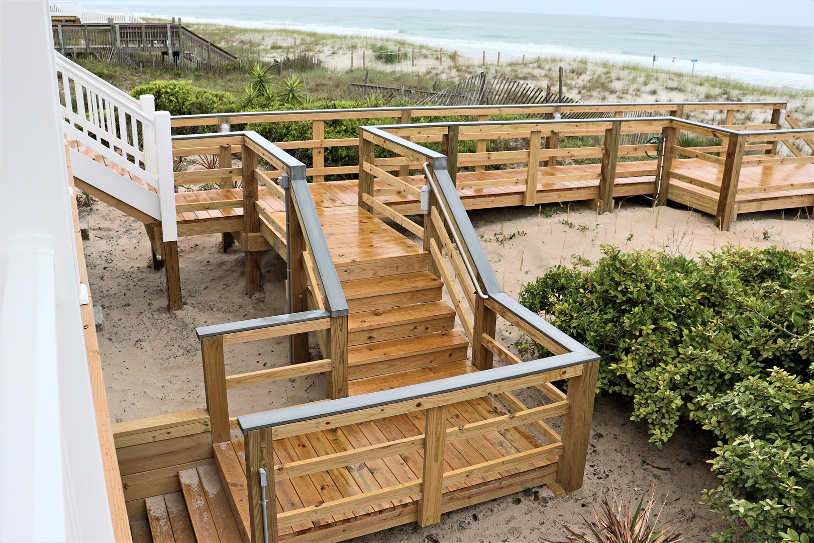 A wooden boardwalk with accessibility features leads through coastal dunes toward the beach and ocean waves.