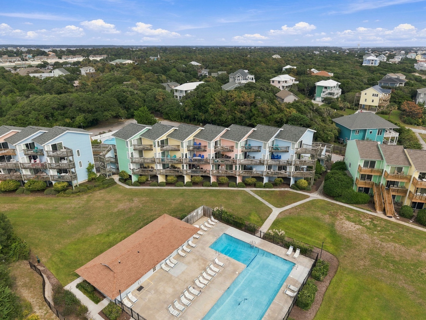 Aerial view of a coastal vacation community featuring colorful townhomes, a shared swimming pool, and abundant natural surroundings with maritime forest and neighboring beach homes.