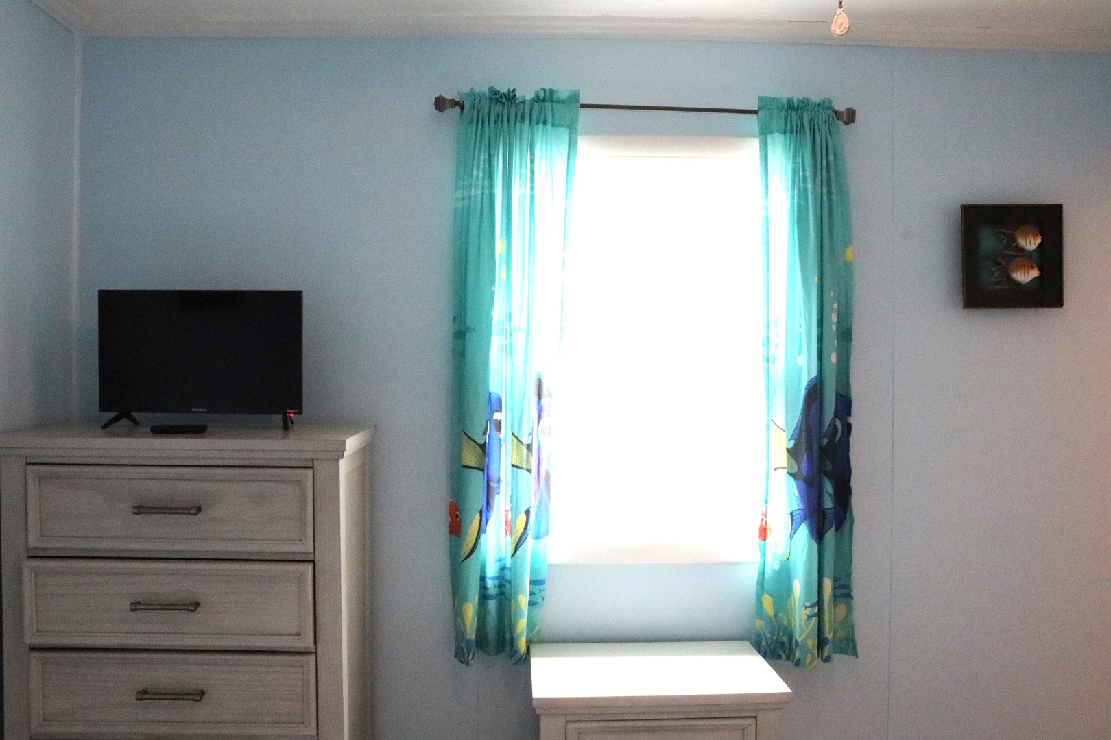 Your bedroom is bathed in natural light through cheerful turquoise curtains with playful fish designs, complemented by a mounted TV and coastal-themed artwork.