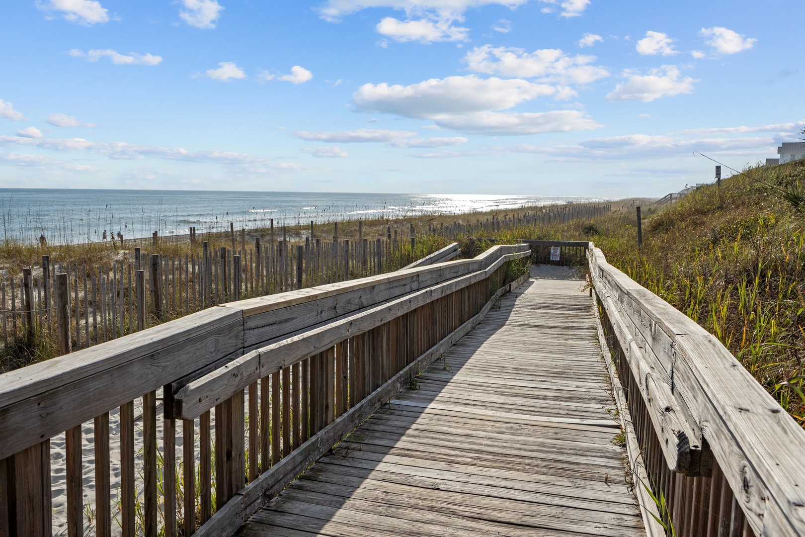 Wooden boardwalk leads through coastal dunes to pristine sandy beach and ocean waves under bright blue sky.
